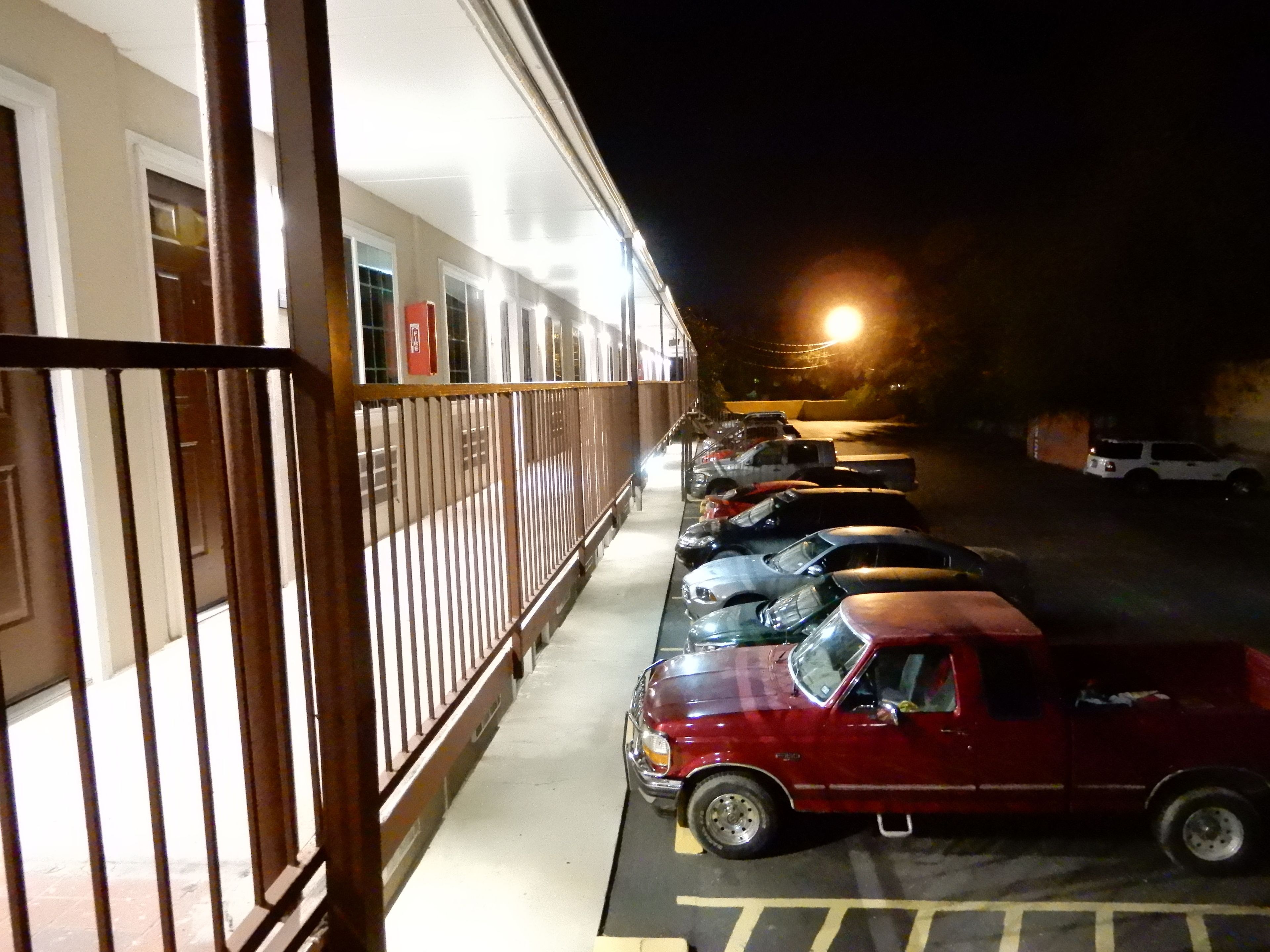 Un parking où plusieurs voitures sont garées à la suite, un camion rouge et, au fond, un immeuble doté d’un balcon.