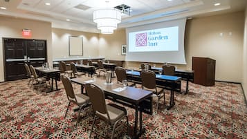Ein Konferenzraum mit floralem Teppich, einer Projektionsleinwand, die Hilton Garden Inn zeigt, und einem Rednerpult.