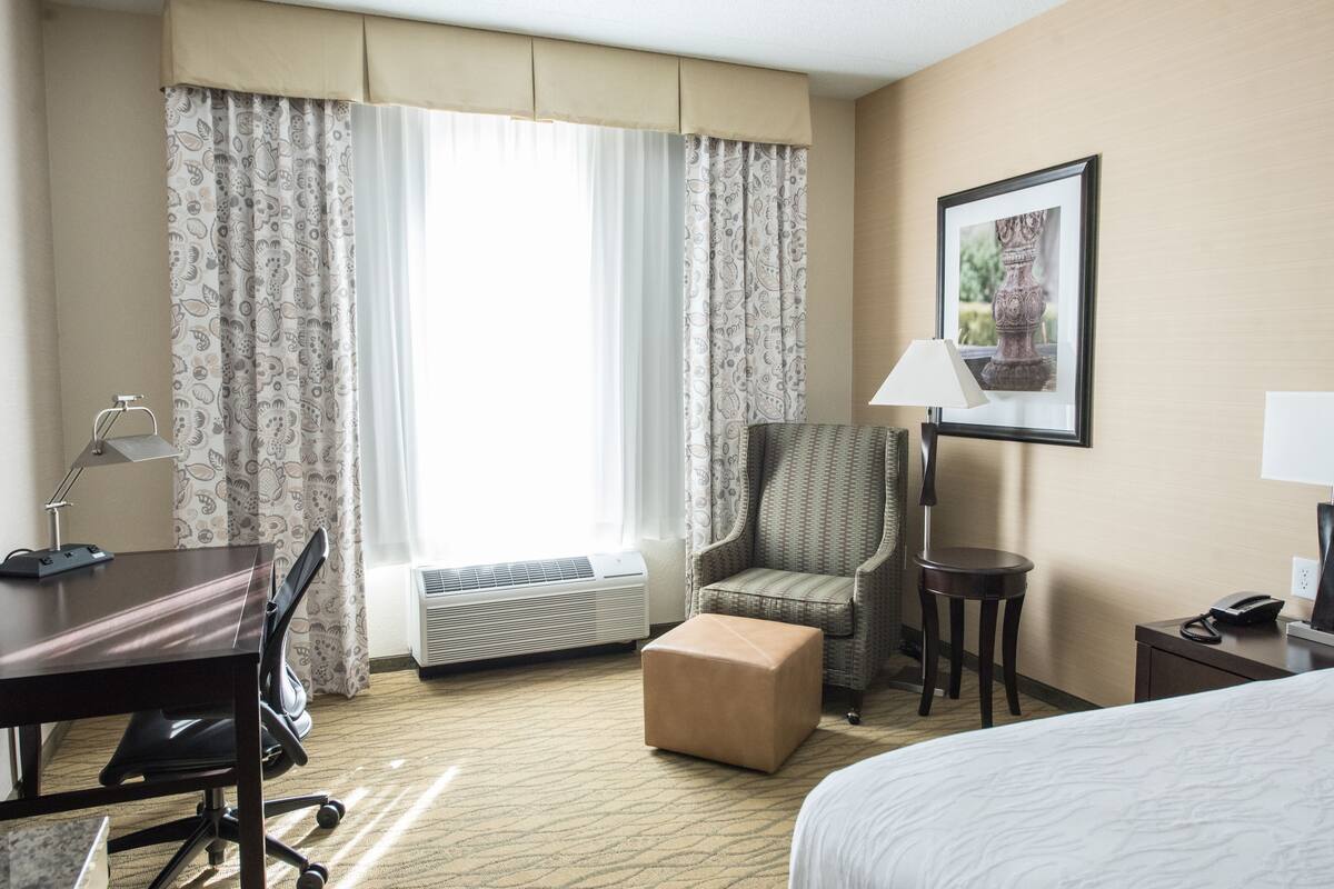 A hotel room with a bed, a desk with a chair, an air conditioner, a small ottoman, a lamp, and a framed picture on the wall.