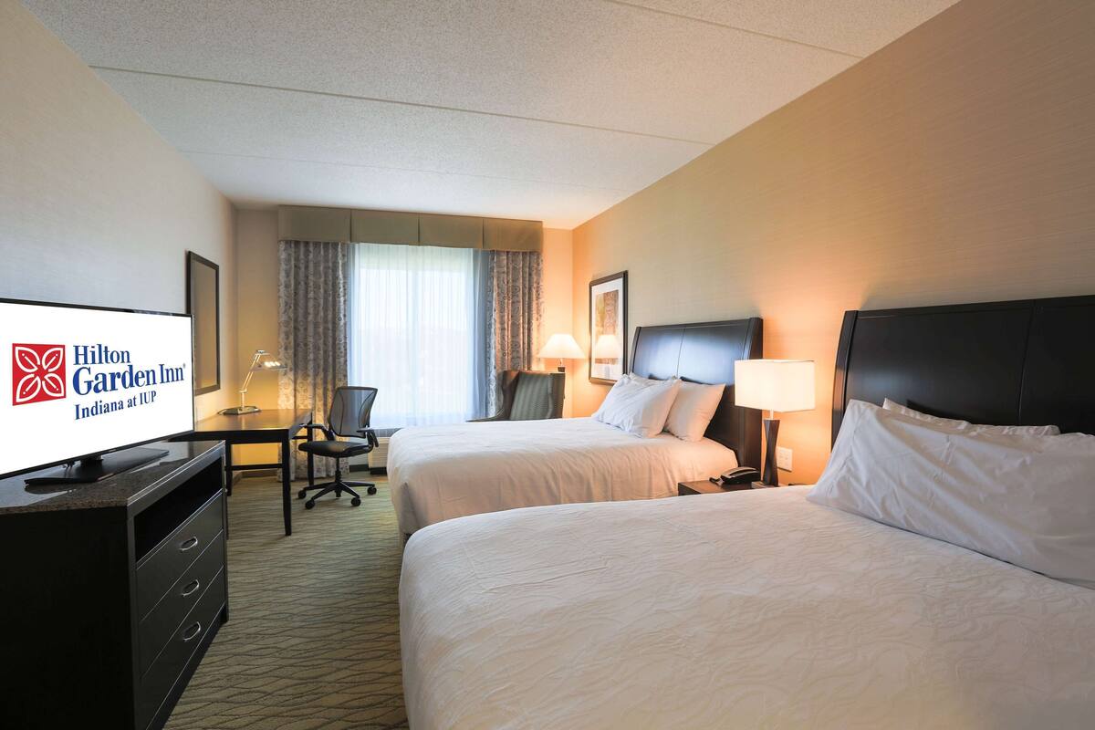 A hotel room with two beds, a desk, a chair, a television, and a lamp.