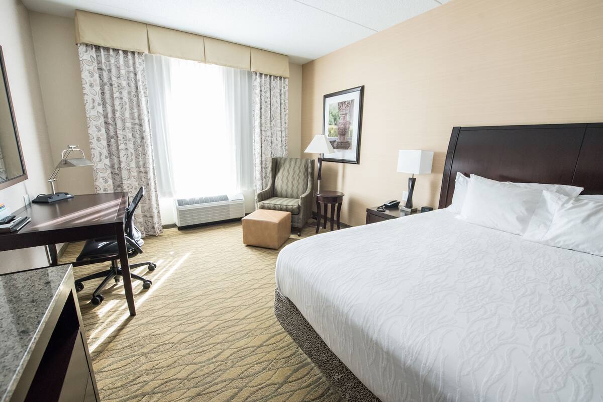 A hotel room with a large bed, a desk with a chair, a small table, a lamp, and a chair.