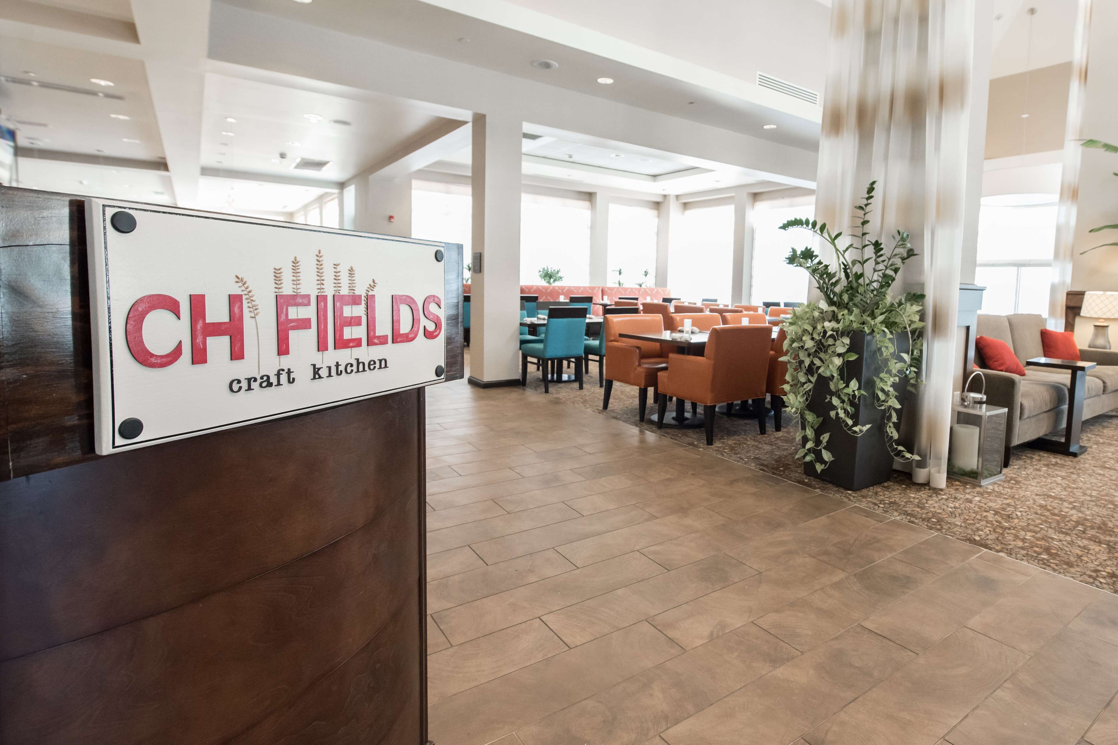 A spacious dining area with multiple tables and chairs, a sign for 'CH FIELDS Craft Kitchen', and a modern interior design with large windows and decorative plants.