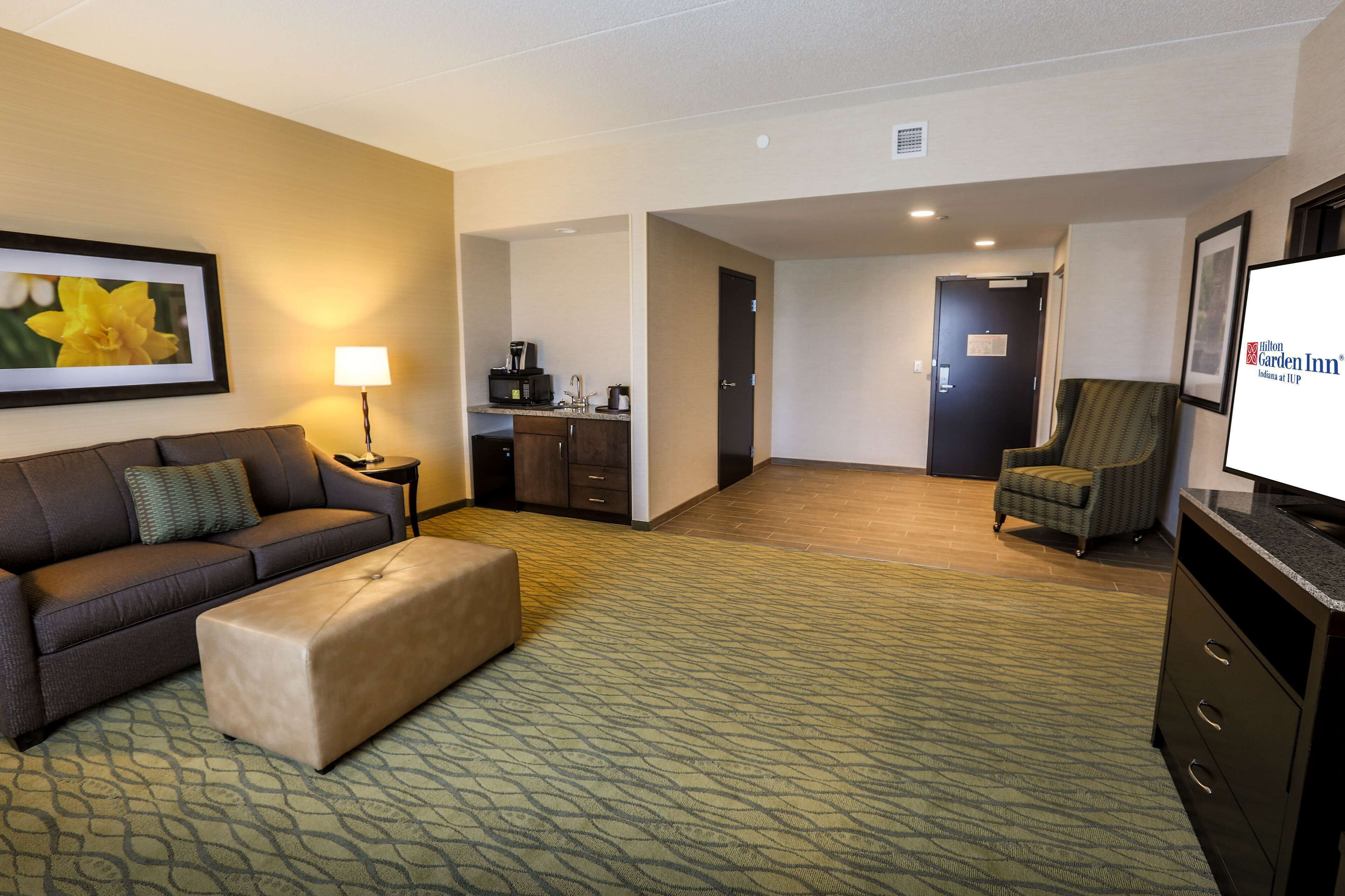 A hotel room with a sofa, ottoman, a chair, a television, a desk with a lamp, and a kitchenette.