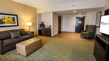 A hotel room with a sofa, ottoman, a chair, a television, a desk with a lamp, and a kitchenette.