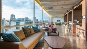 Un hall d’hôtel moderne offrant une vue imprenable sur la ville, avec un canapé agrémenté de coussins à motifs et une table basse en bois.