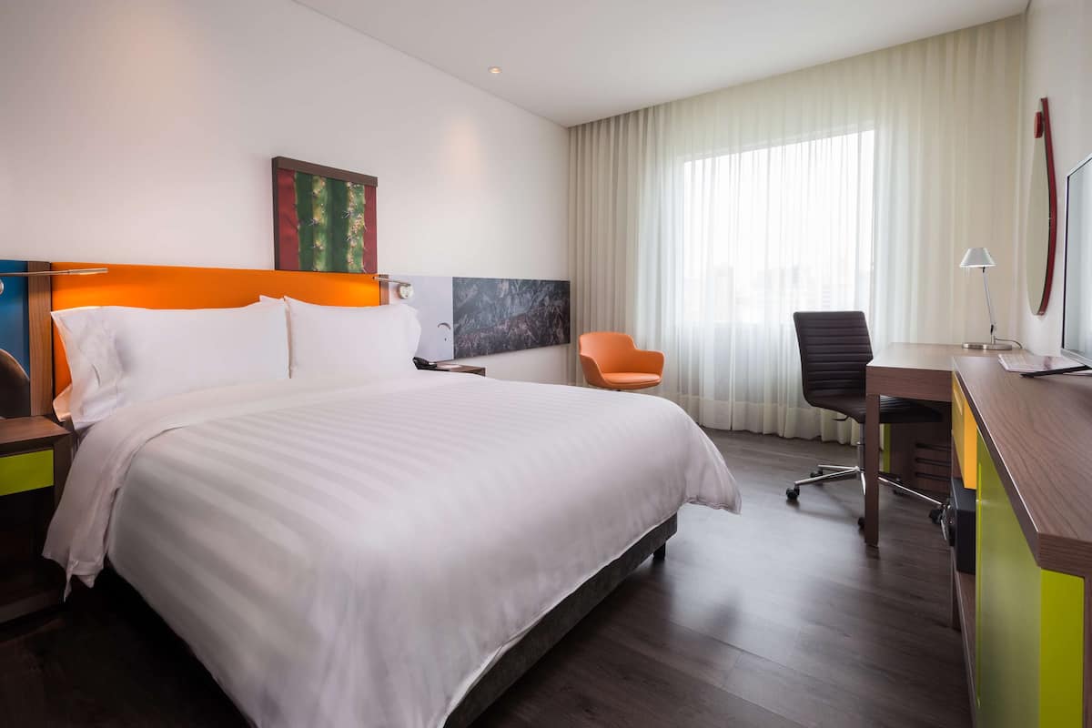 Une chambre d’hôtel moderne avec un grand lit, un bureau, un fauteuil orange et une grande fenêtre avec des rideaux.