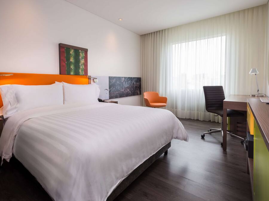 Una habitación de hotel moderna con una cama grande, un escritorio, una silla naranja y un ventanal con cortinas.