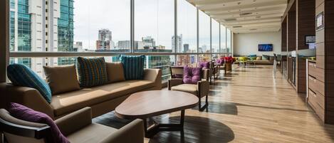 Un área de descanso moderna con sofá, sillas y una mesa de centro. Amplios ventanales ofrecen vistas a la ciudad.