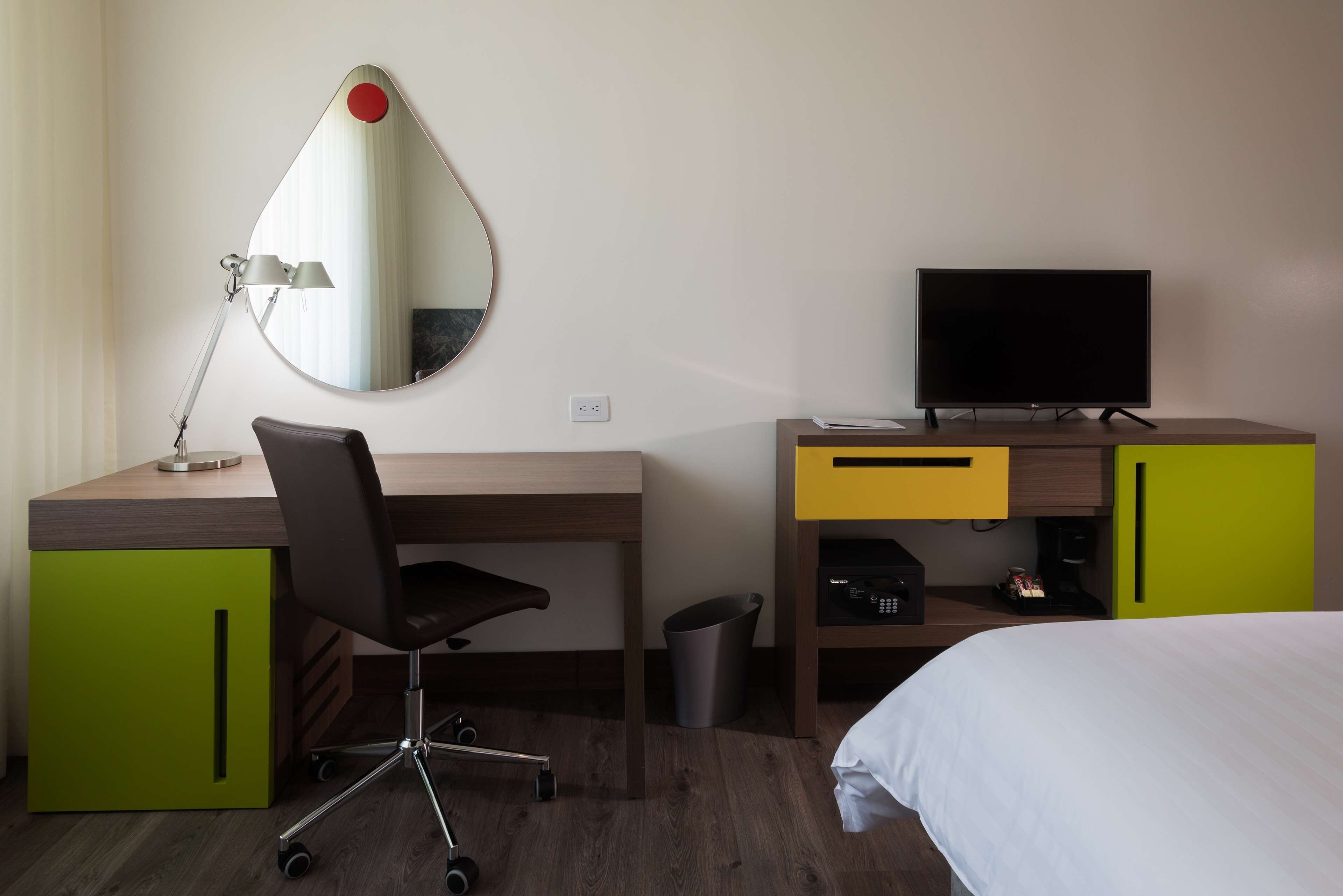 A hotel room with a wooden desk, a chair, a television, a bed, and a mirror.