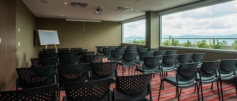Un salón de conferencias con filas de sillas negras, una pizarra y un amplio ventanal con vistas al mar y a las montañas.