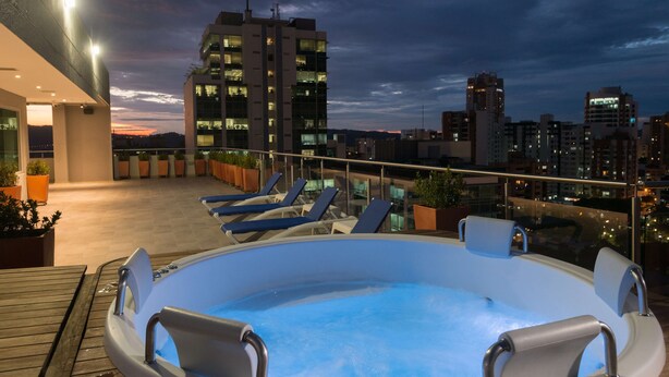 Un espace piscine aménagé sur le toit, avec des chaises longues et un jacuzzi, offrant une vue sur le paysage urbain au crépuscule.