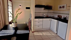 Een kleine keuken met witte kastjes, een zwart aanrechtblad en een magnetron. Op de tafel staat een plant in pot en aan de muur hangt een lijst.