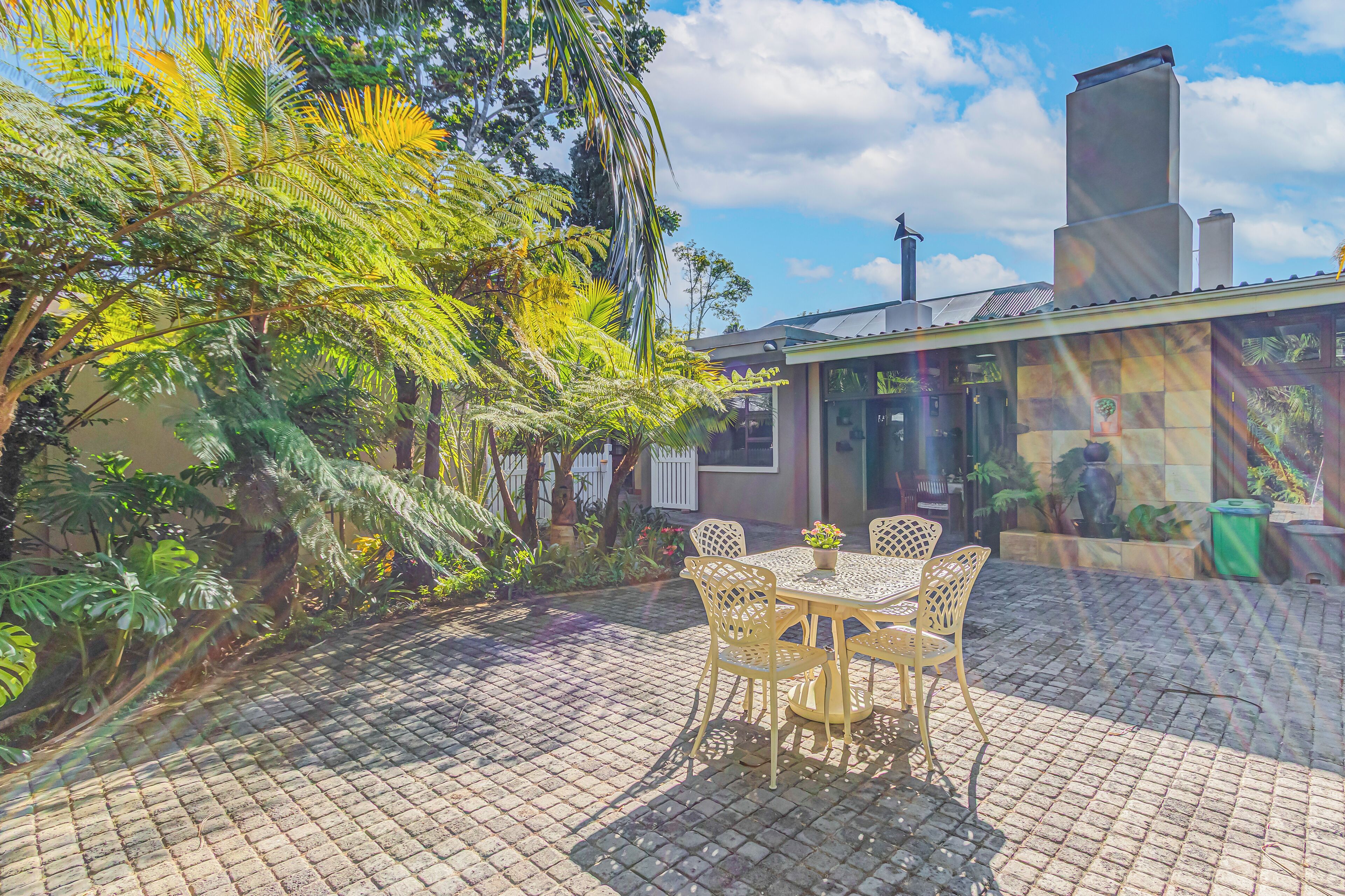 A patio with a table and chairs, surrounded by lush greenery and a building with a chimney.