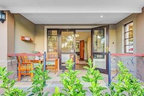 A hotel entrance with a wooden bench and chairs outside, a tiled patio, and a metal railing.