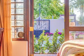 A room with a wooden door, a window with a metal grill, and a view of a garden with plants and a fence.