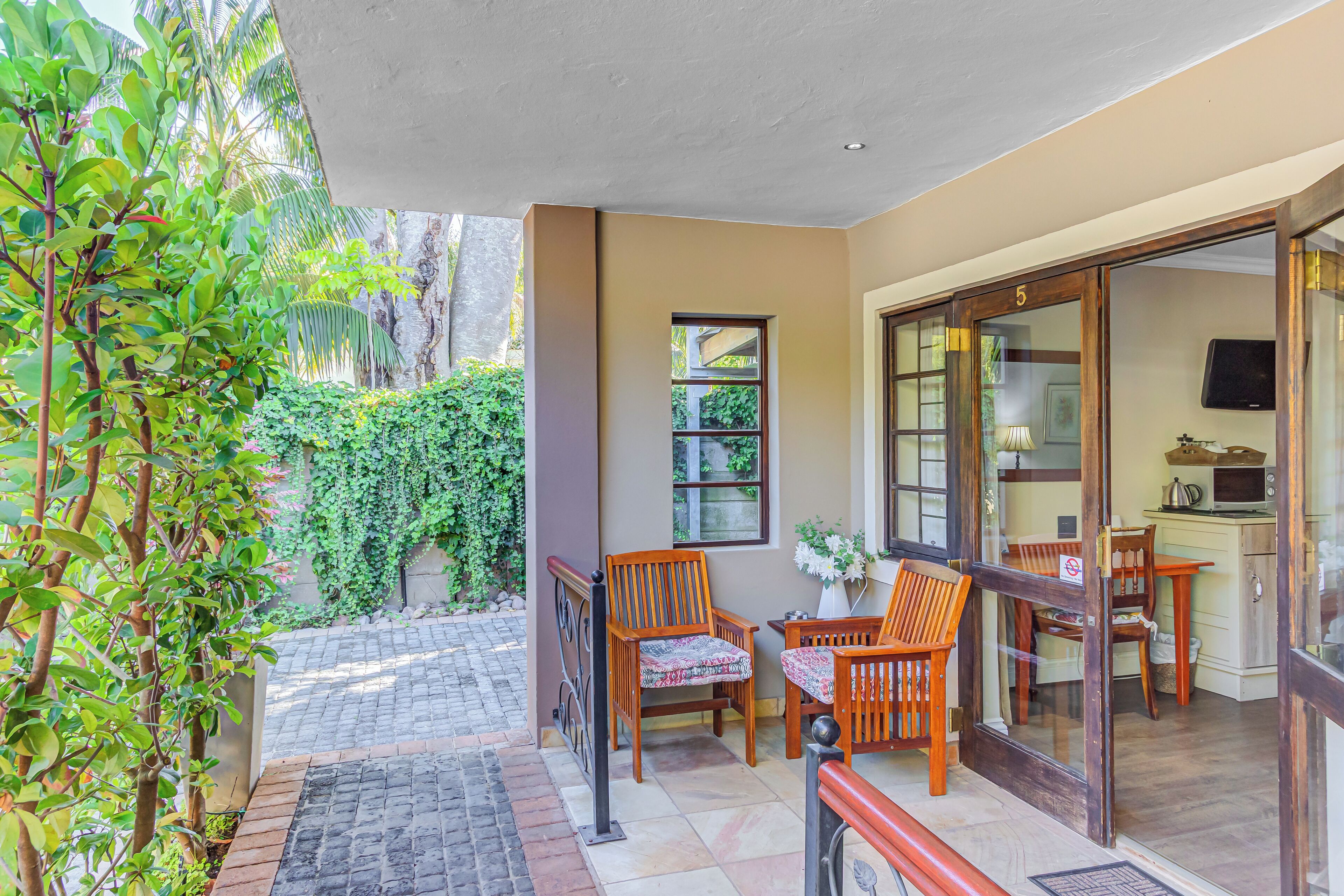 A patio area with a wooden table and chairs, a small kitchen area, and a view of a garden with greenery.