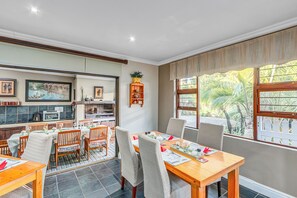 A dining area with a wooden table and chairs, a kitchen in the background, and large windows with a view of palm trees.