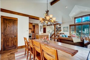 A spacious dining area with a wooden table and chairs, a chandelier, and a view of the kitchen and living room in the background.