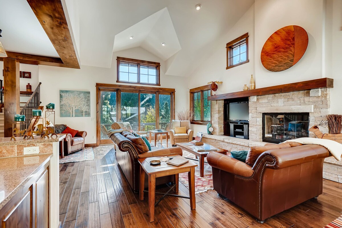A spacious living room with a fireplace, wooden floors, and large windows offering a view of the outdoors.