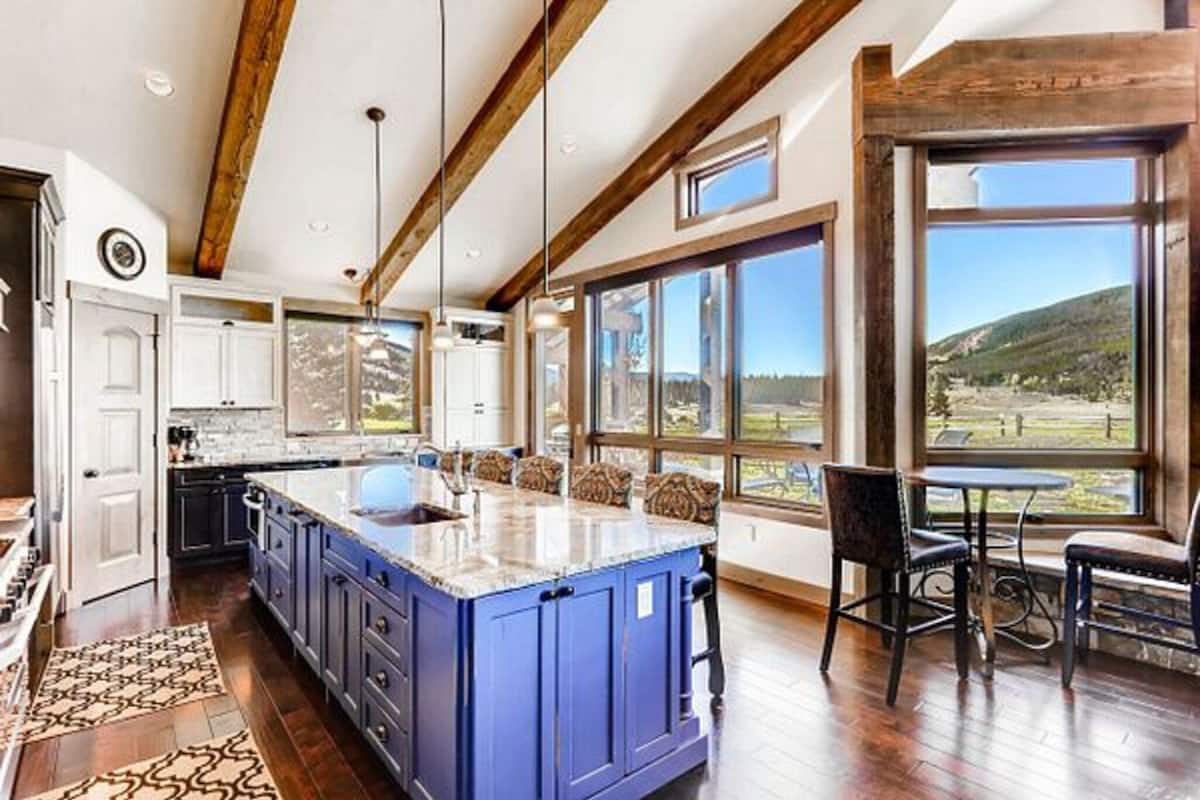 A kitchen with a blue island, wooden beams, large windows, and a view of the outdoors.