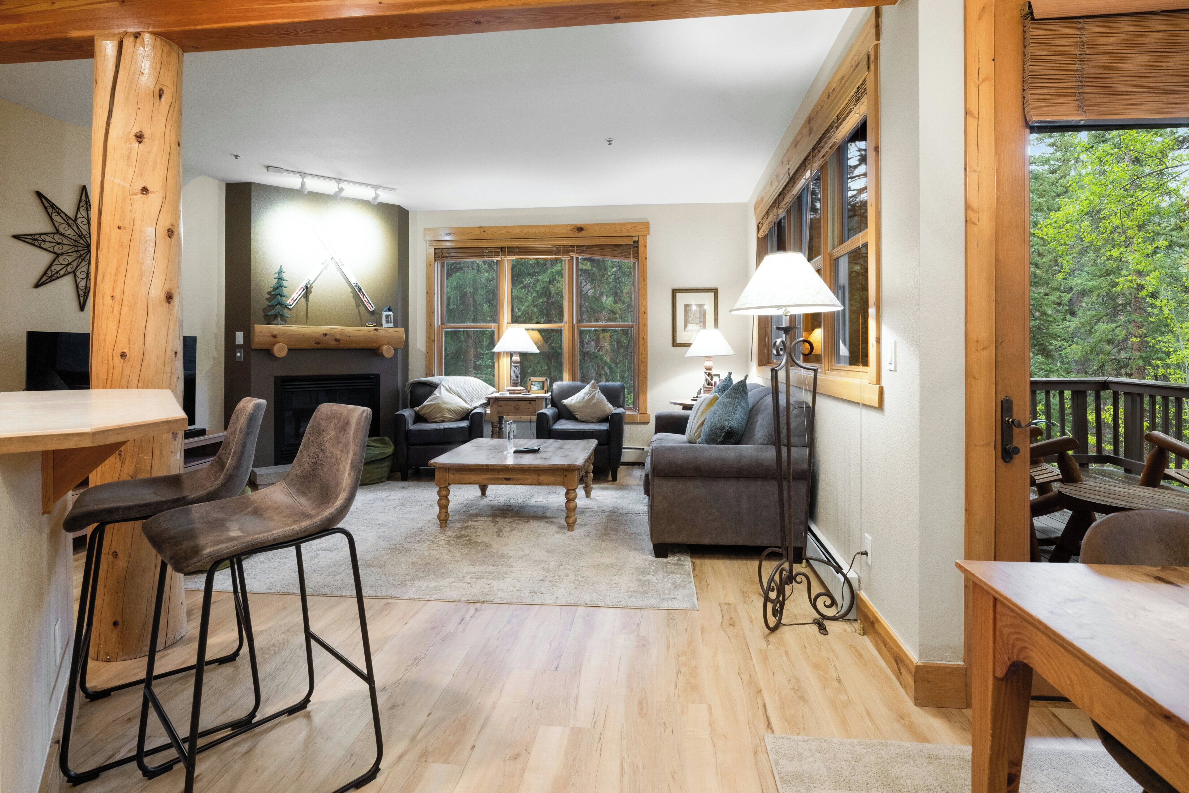 A cozy living room with a fireplace, wooden furniture, and large windows offering a view of the outdoors.