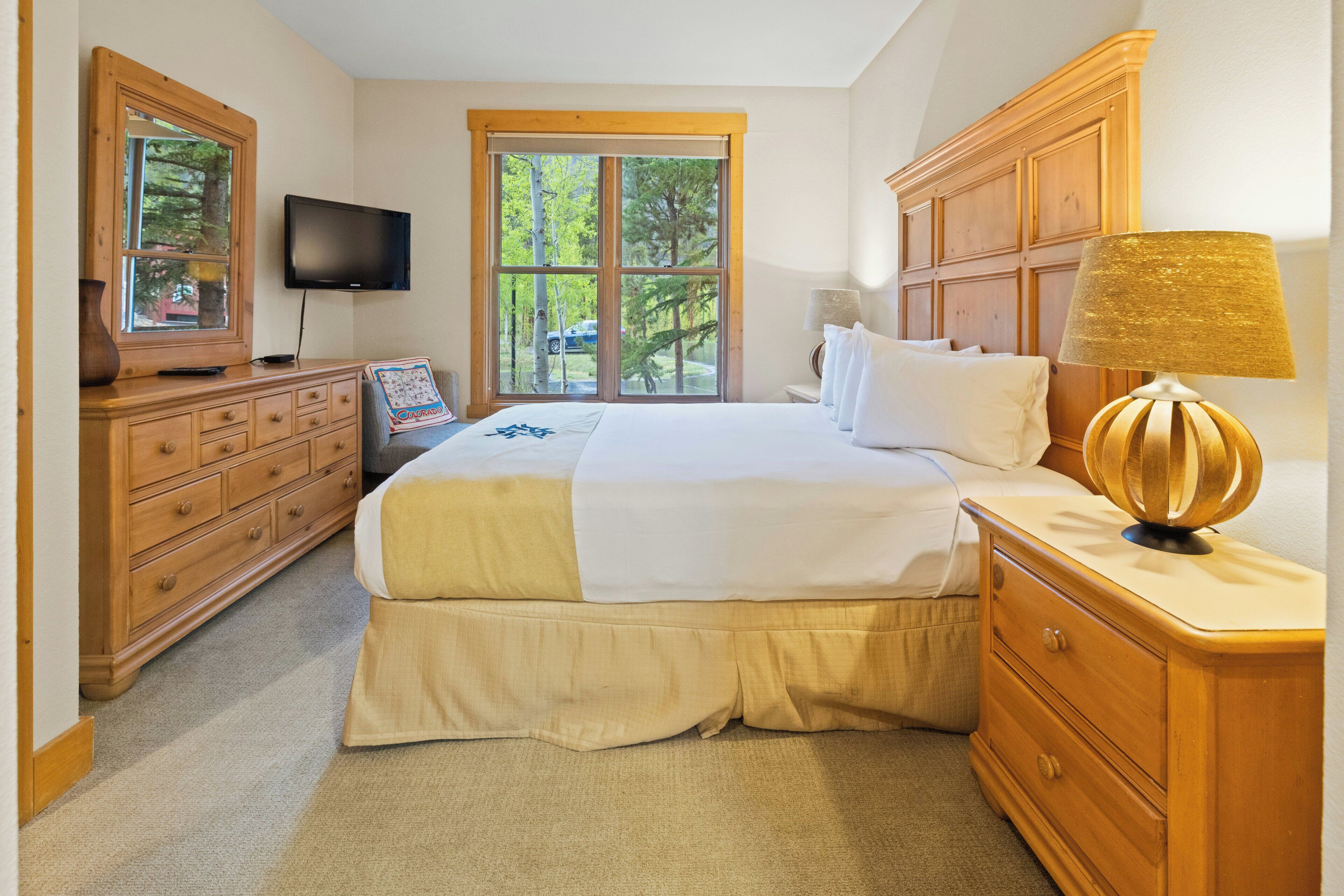 A bedroom with a large bed, wooden dresser, a television, and a window with a view of trees.