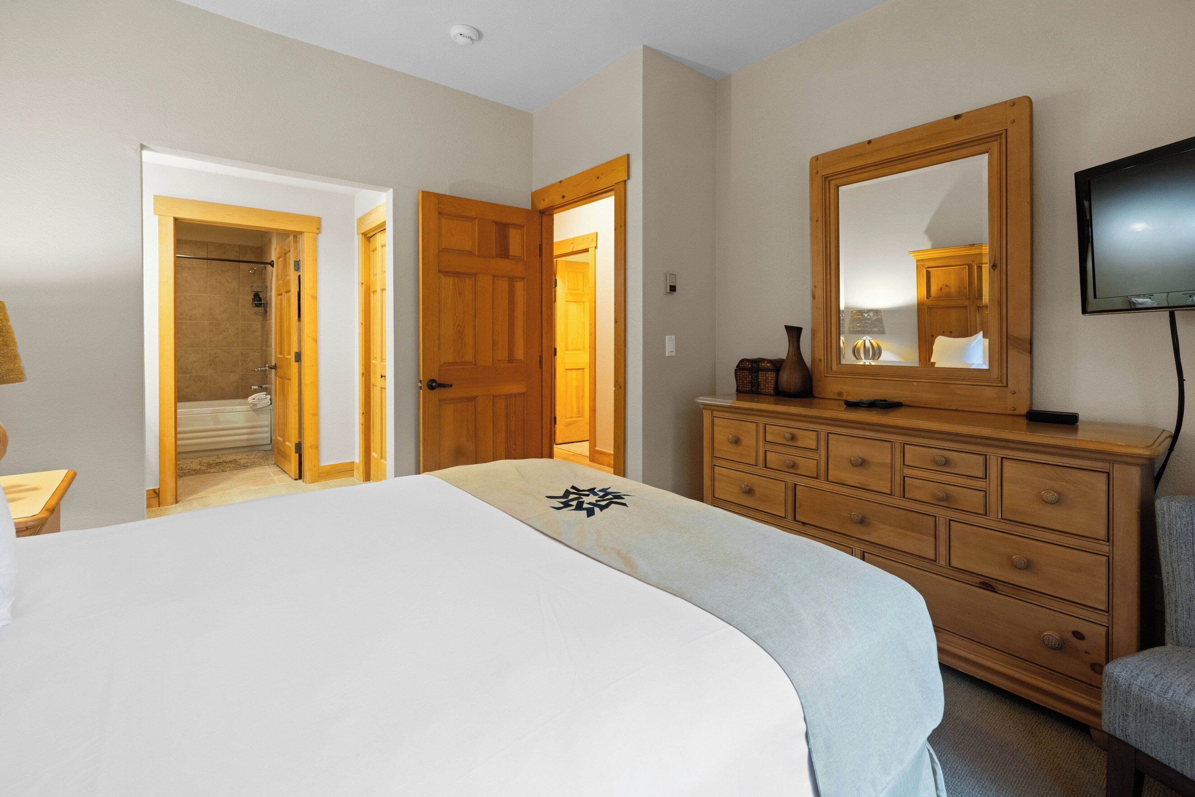 A bedroom with a large bed, wooden dresser, and a TV mounted on the wall.