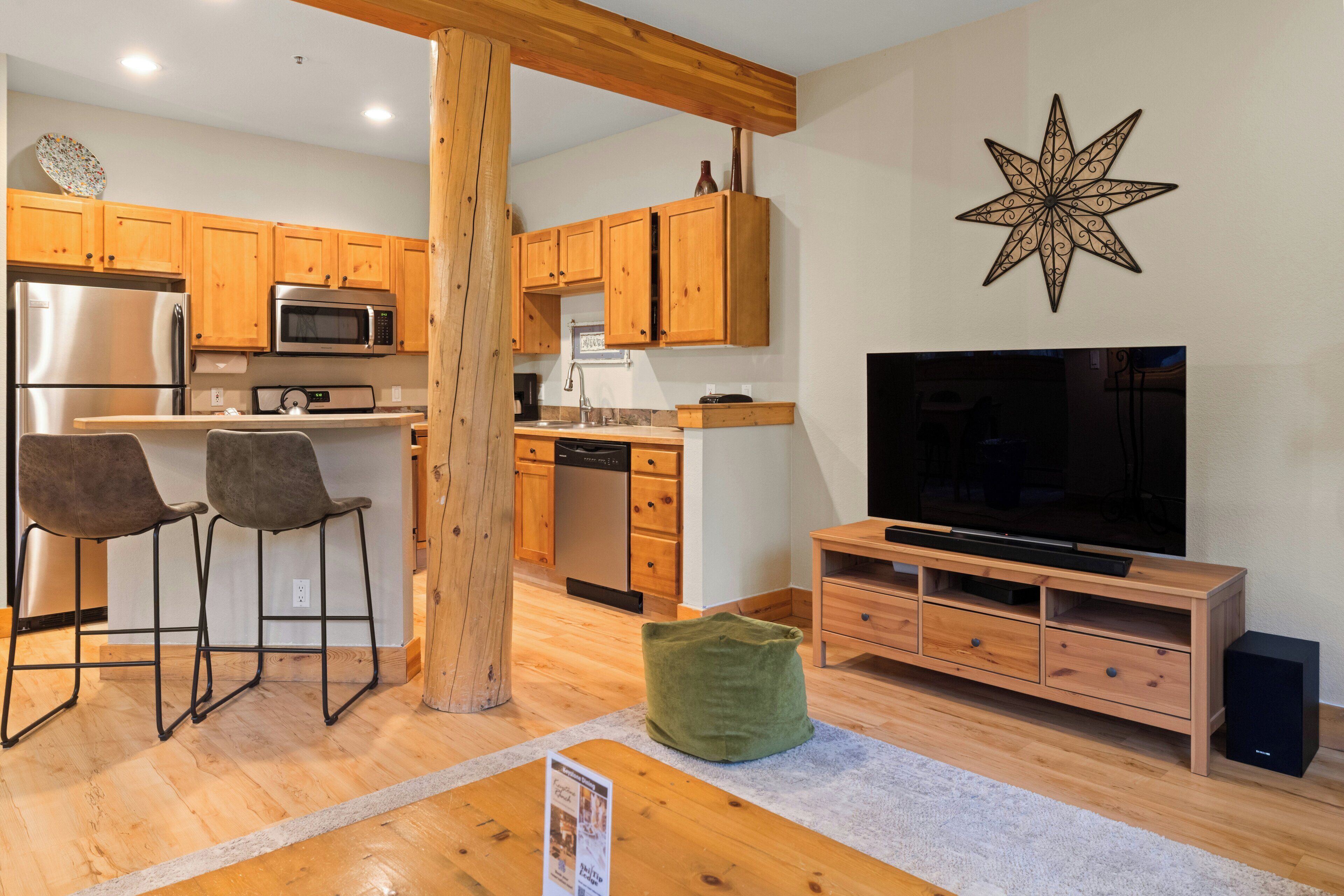 A modern kitchen with wooden cabinets, a microwave, and a refrigerator. There is a dining area with two chairs and a table. A television is on a wooden stand, and a star-shaped decoration is on the wall.