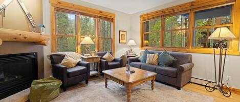 A cozy living room with a fireplace, two sofas, a coffee table, and large windows with wooden frames.