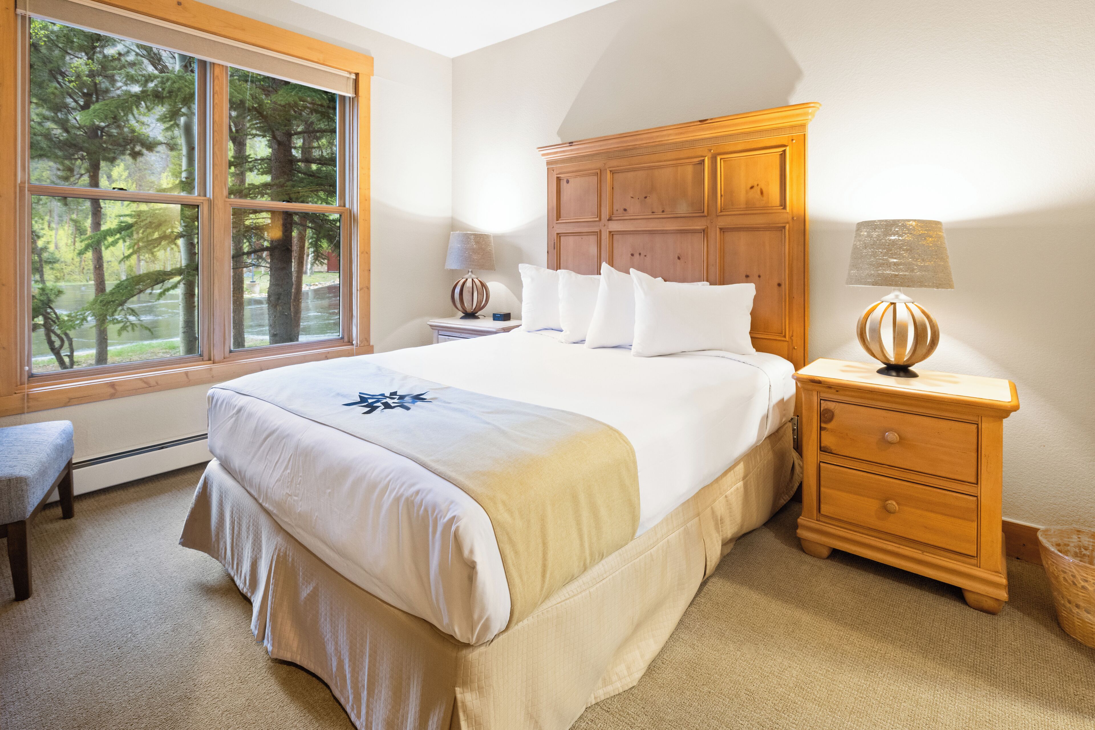Une chambre à coucher avec un grand lit, une table de chevet en bois, une lampe et une vue sur les arbres par la fenêtre.