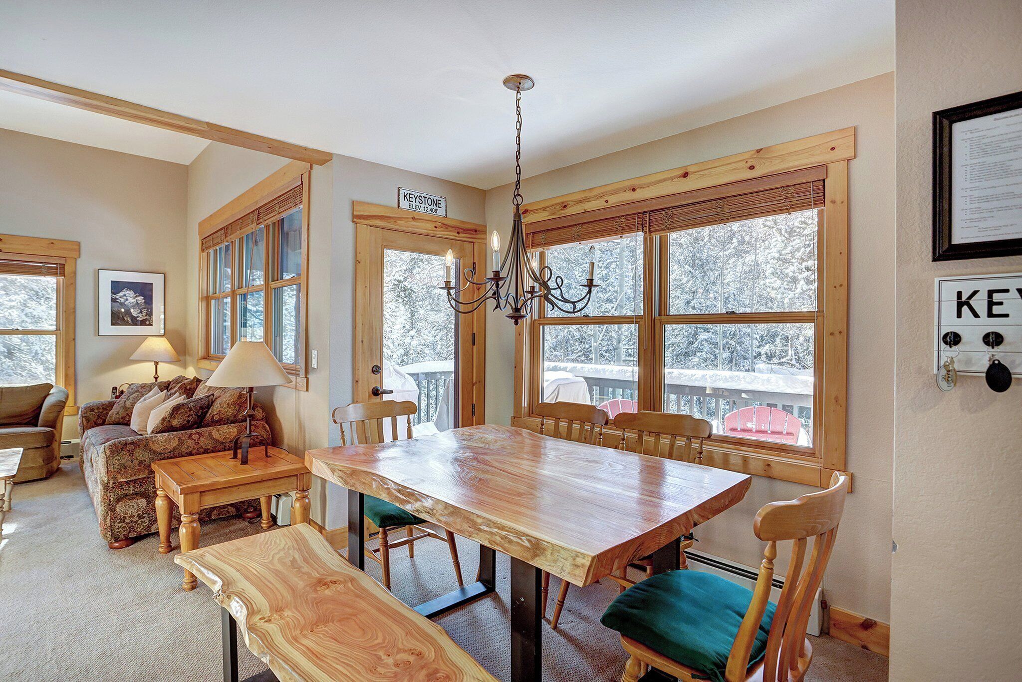 A cozy room with a wooden dining table, benches, and a chandelier. There is a couch with pillows and a lamp. Large windows with wooden frames offer a view of trees outside.