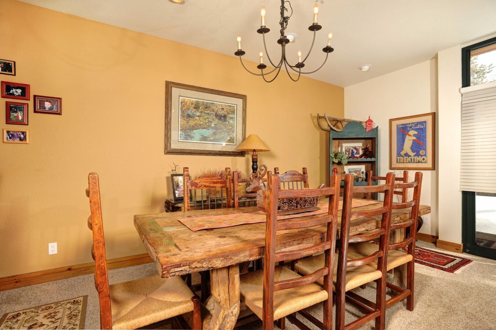 Un coin repas avec une table et des chaises en bois rustique, un lustre, des tableaux encadrés au mur et une fenêtre avec des stores.