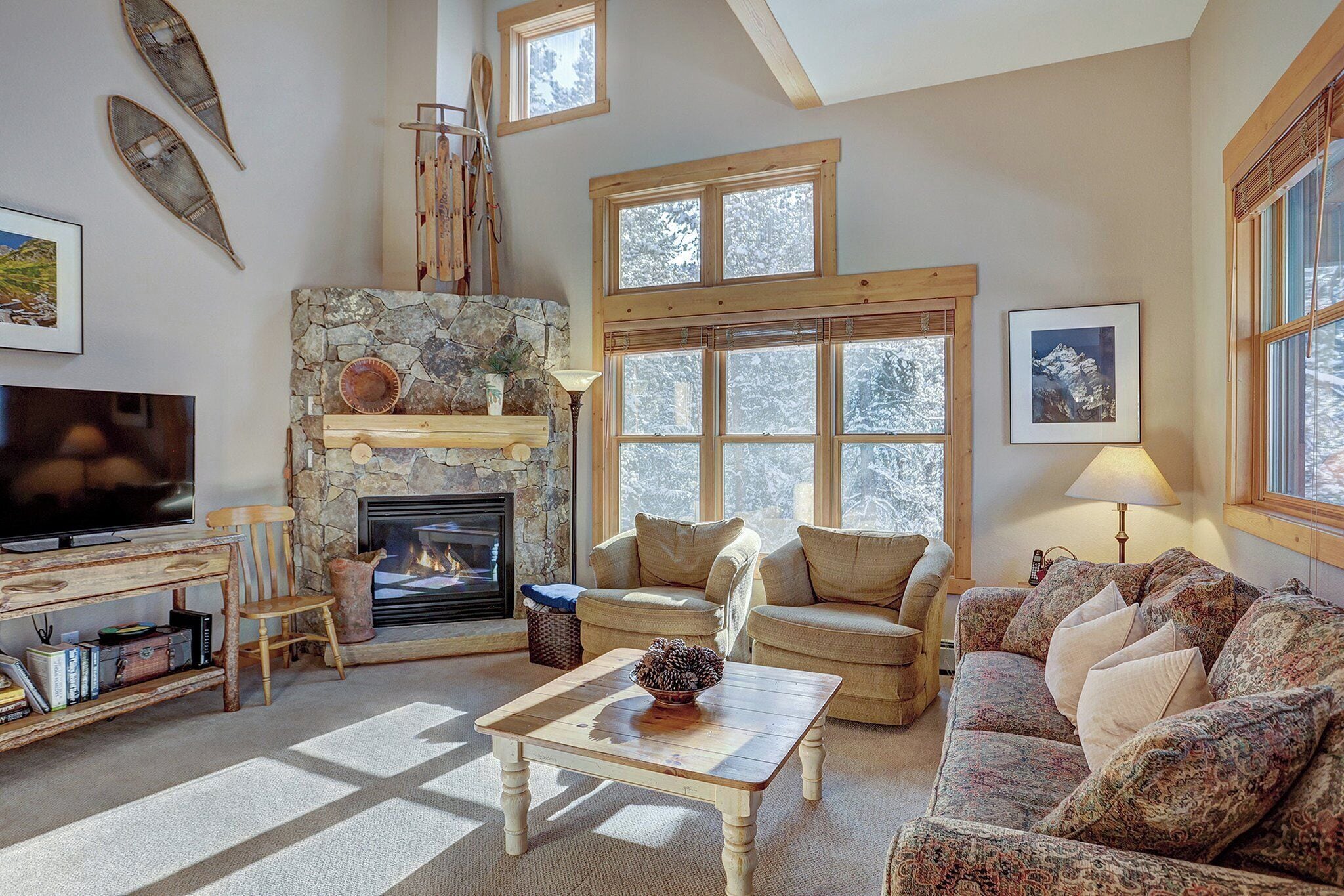 A cozy living room with a stone fireplace, a television, and a large window with a view of snow-covered trees.