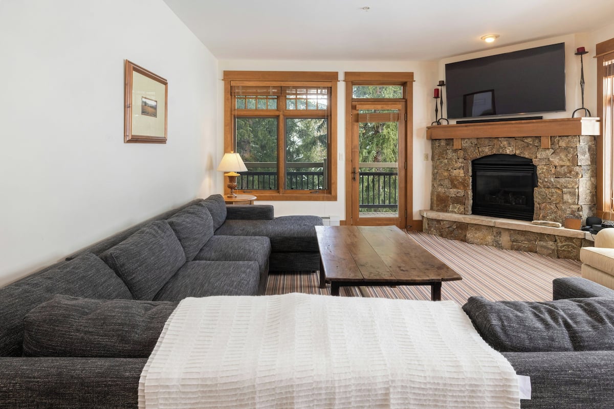 A living room with a sectional sofa, a fireplace, a television mounted above it, and a window with a view of trees.