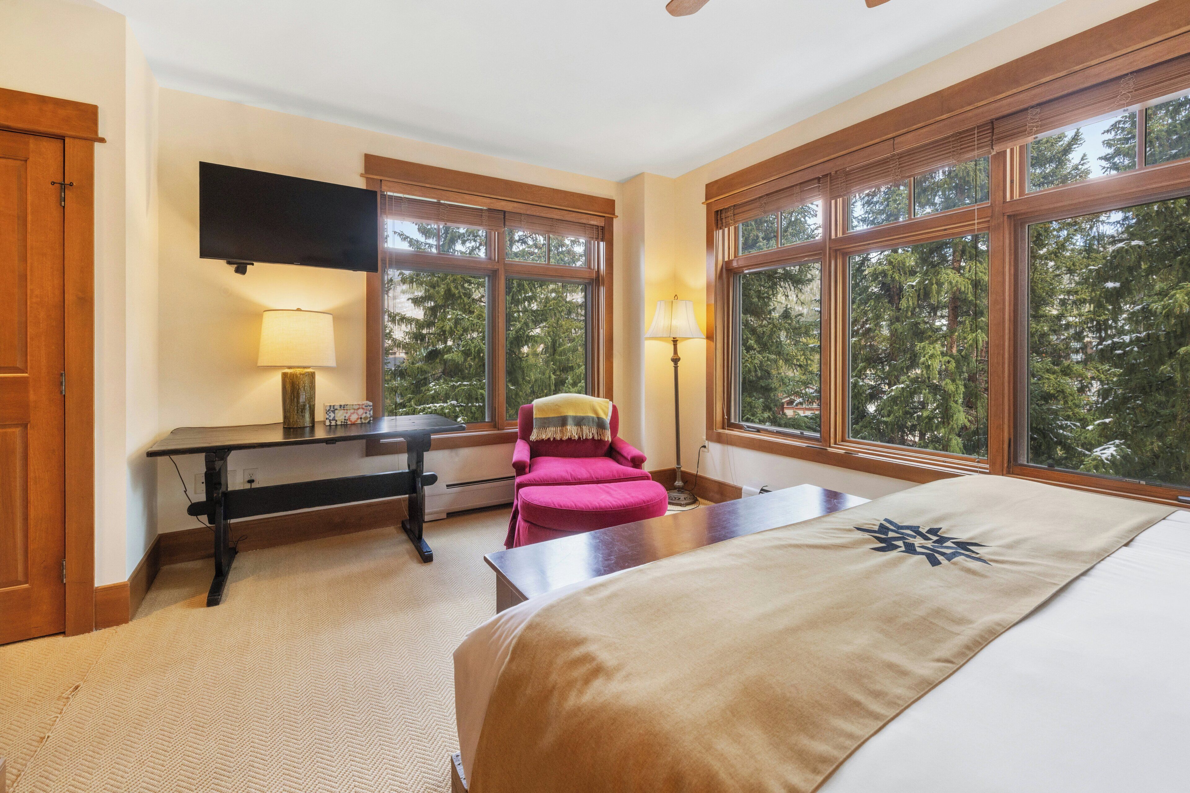 A bedroom with a bed, a desk, a chair, and large windows offering a view of trees.