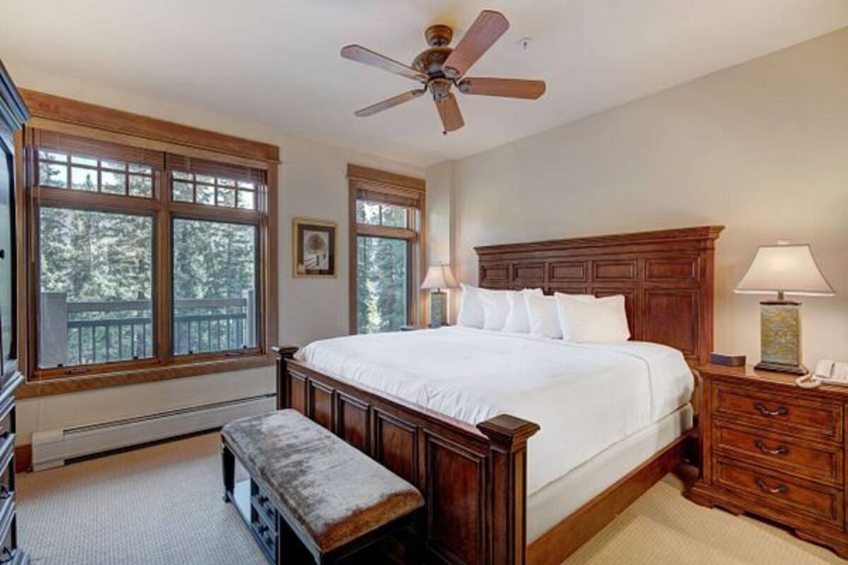 A bedroom with a large bed, wooden headboard, two bedside tables with lamps, a bench, and a ceiling fan.