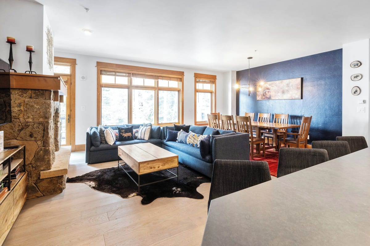 A modern living room with a stone fireplace, a blue sofa, a wooden dining table, and large windows.
