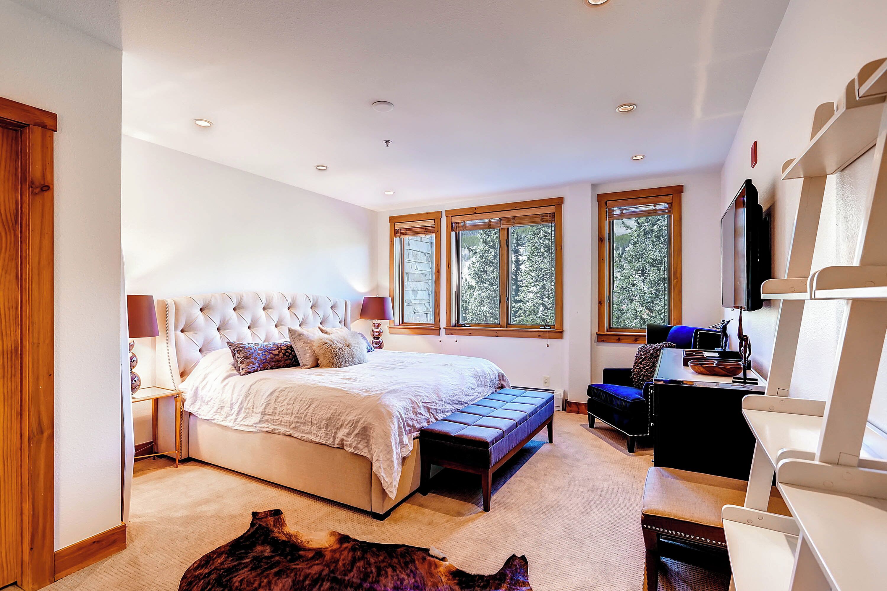 A spacious bedroom with a large bed, a desk, and a chair, a window with a view of trees, and a wooden door.