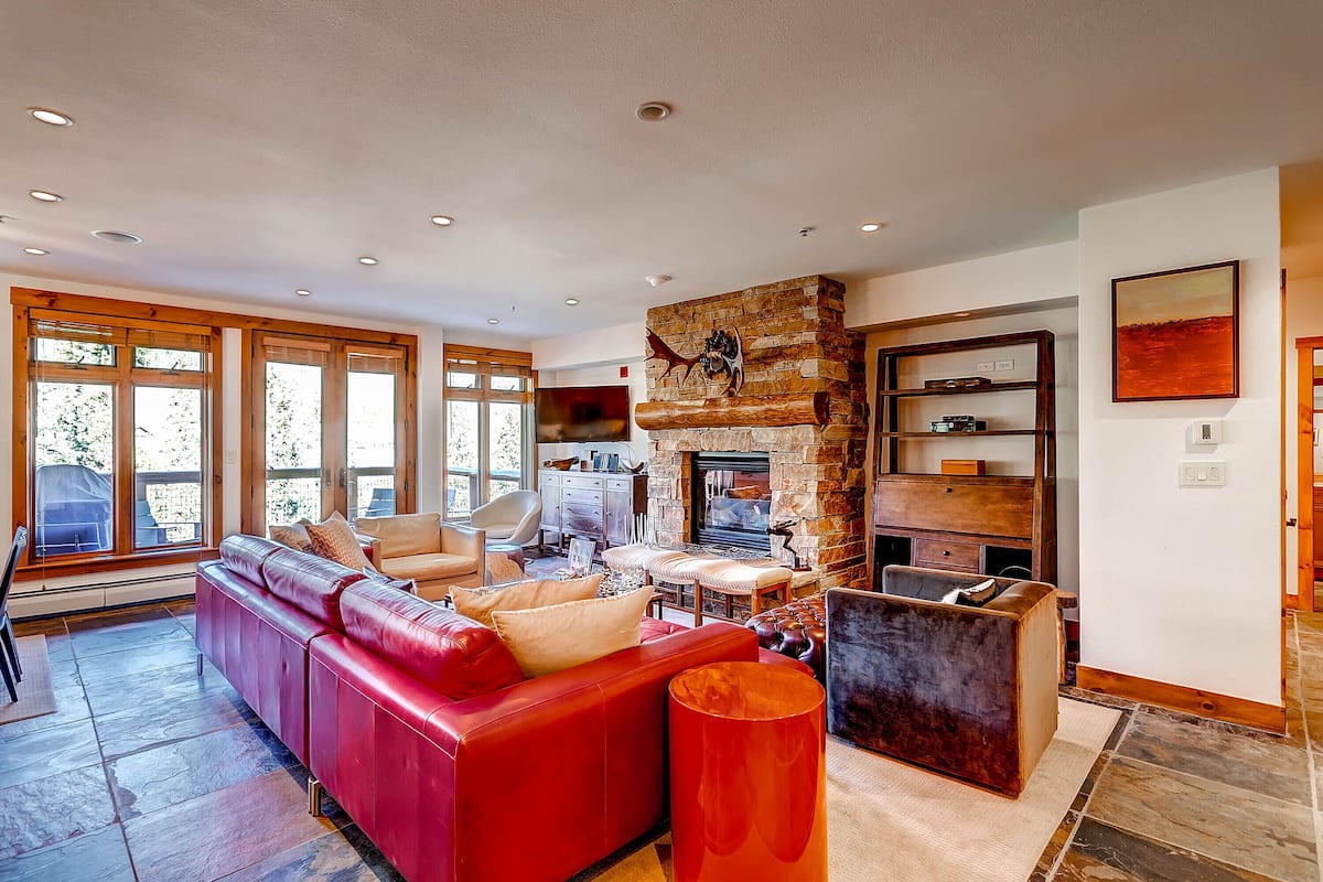 A spacious living room with a red leather sofa, a stone fireplace, and a large window.