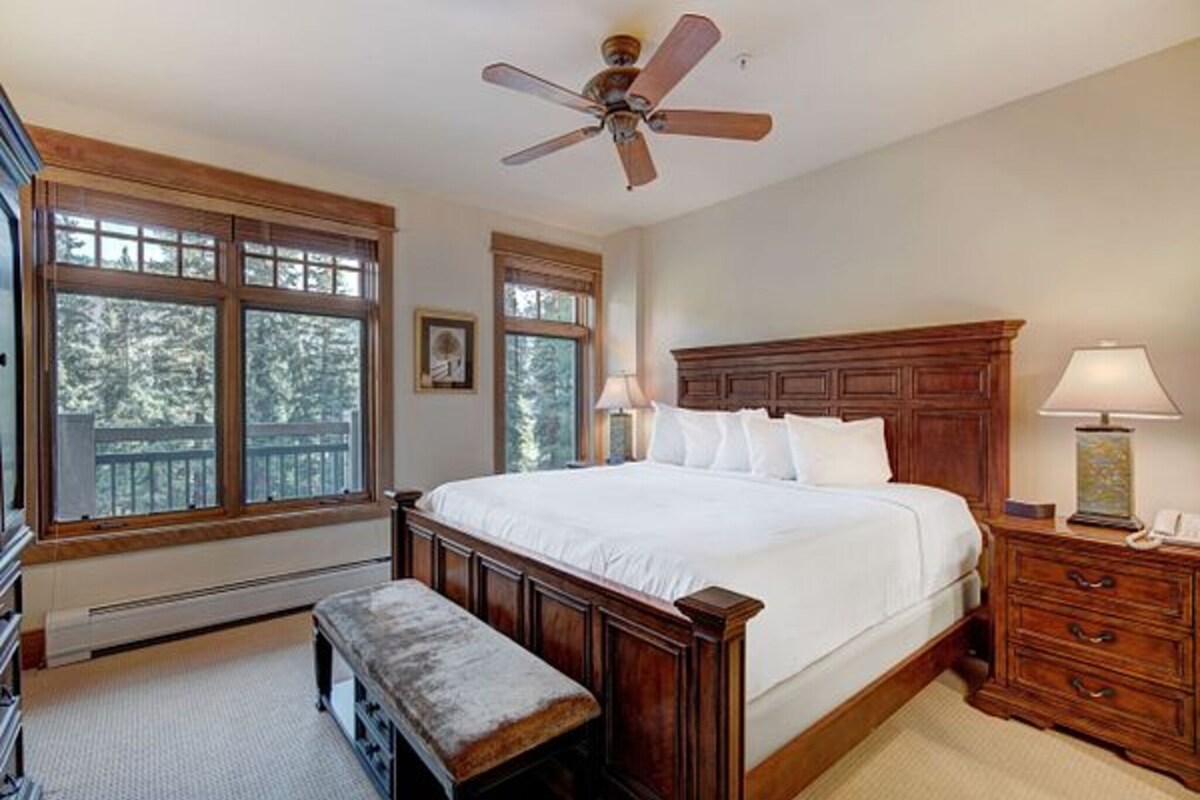A bedroom with a large bed, wooden headboard, two bedside tables with lamps, a bench, and a ceiling fan.