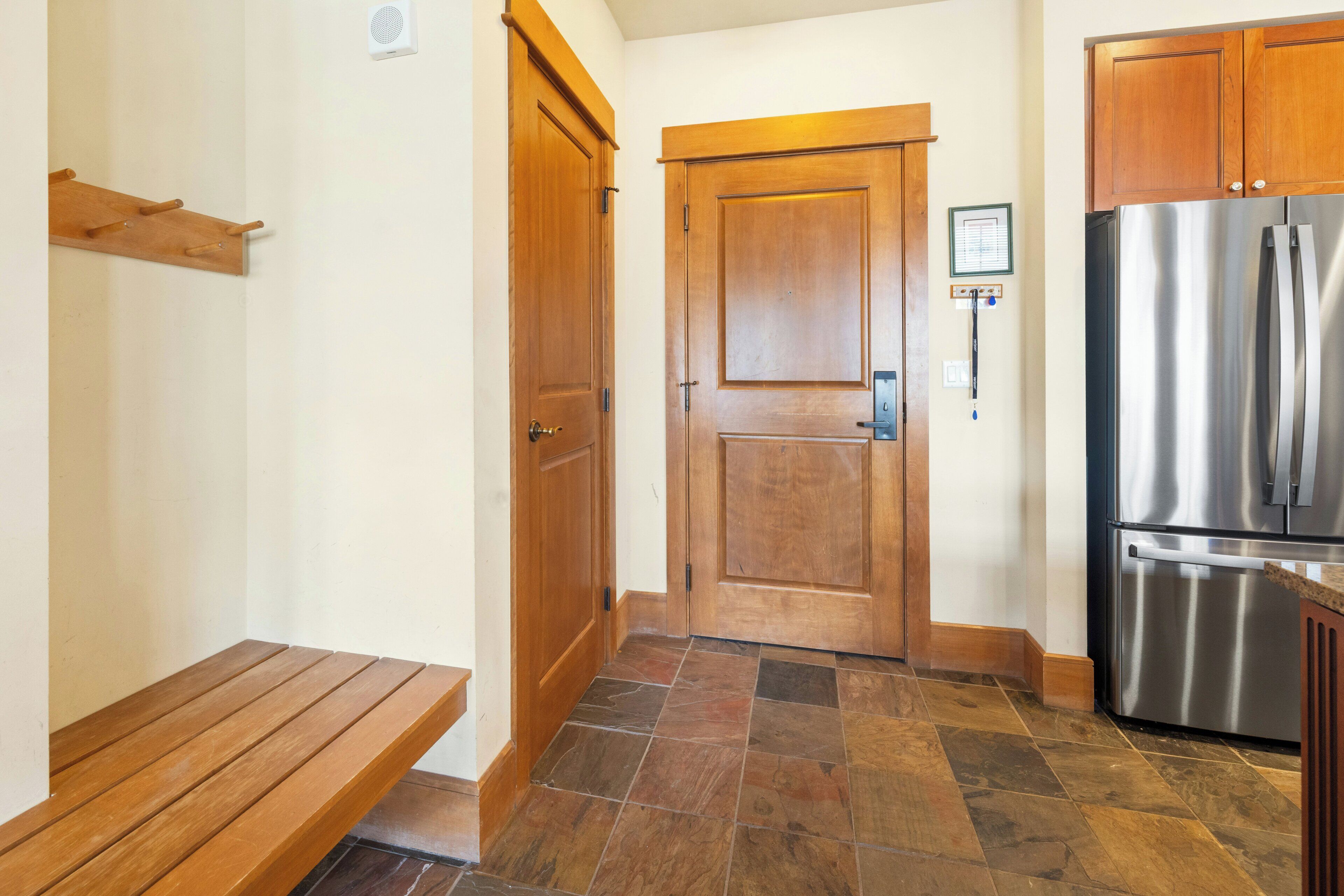 A kitchen with a stainless steel refrigerator, wooden cabinets, a bench, and a tiled floor.