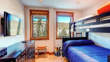 Un dormitorio con literas, un escritorio y una silla, con ventanas que dan a vista de árboles y edificios.