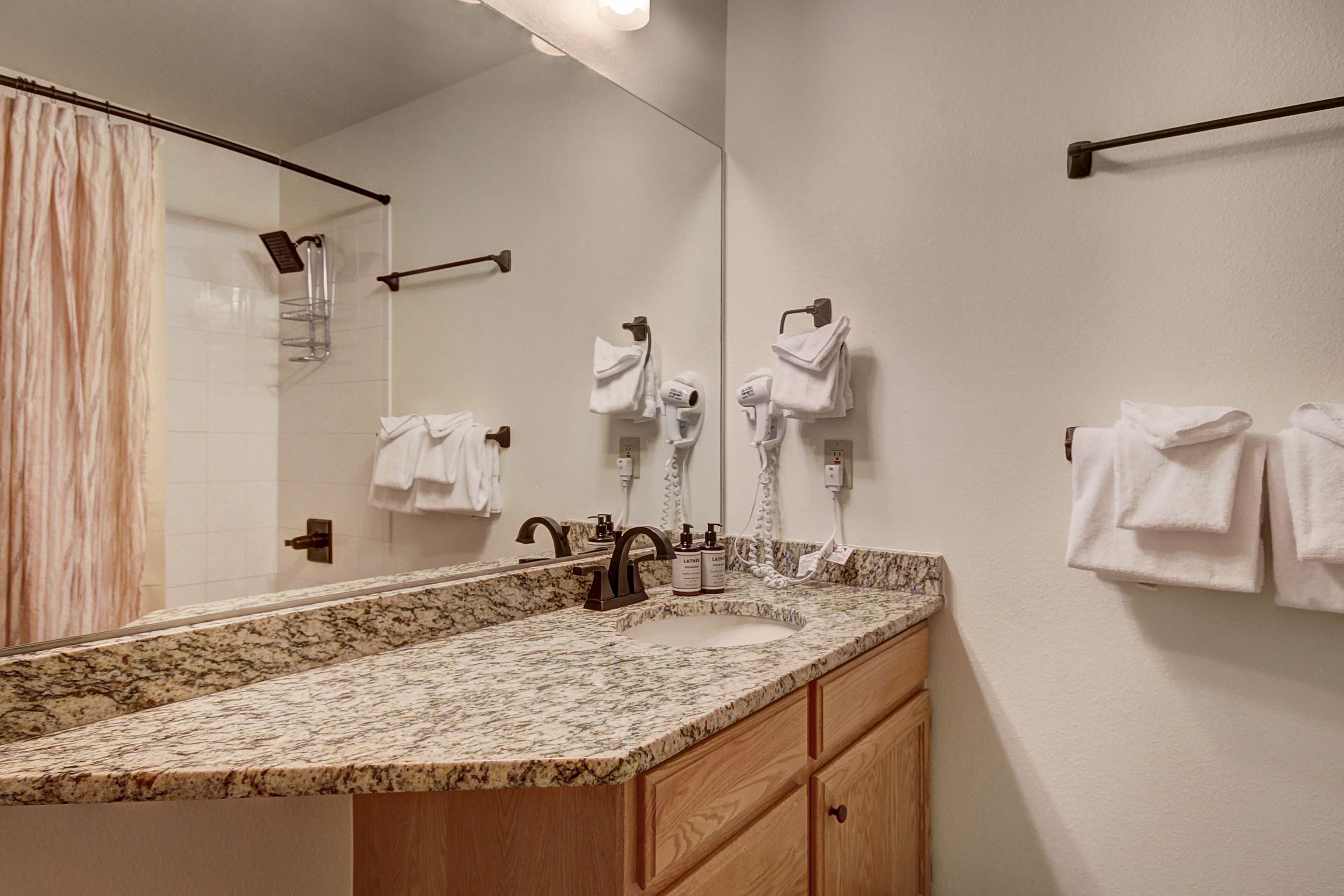A bathroom with a granite countertop sink, wooden cabinets, a large mirror, and multiple towel hooks.