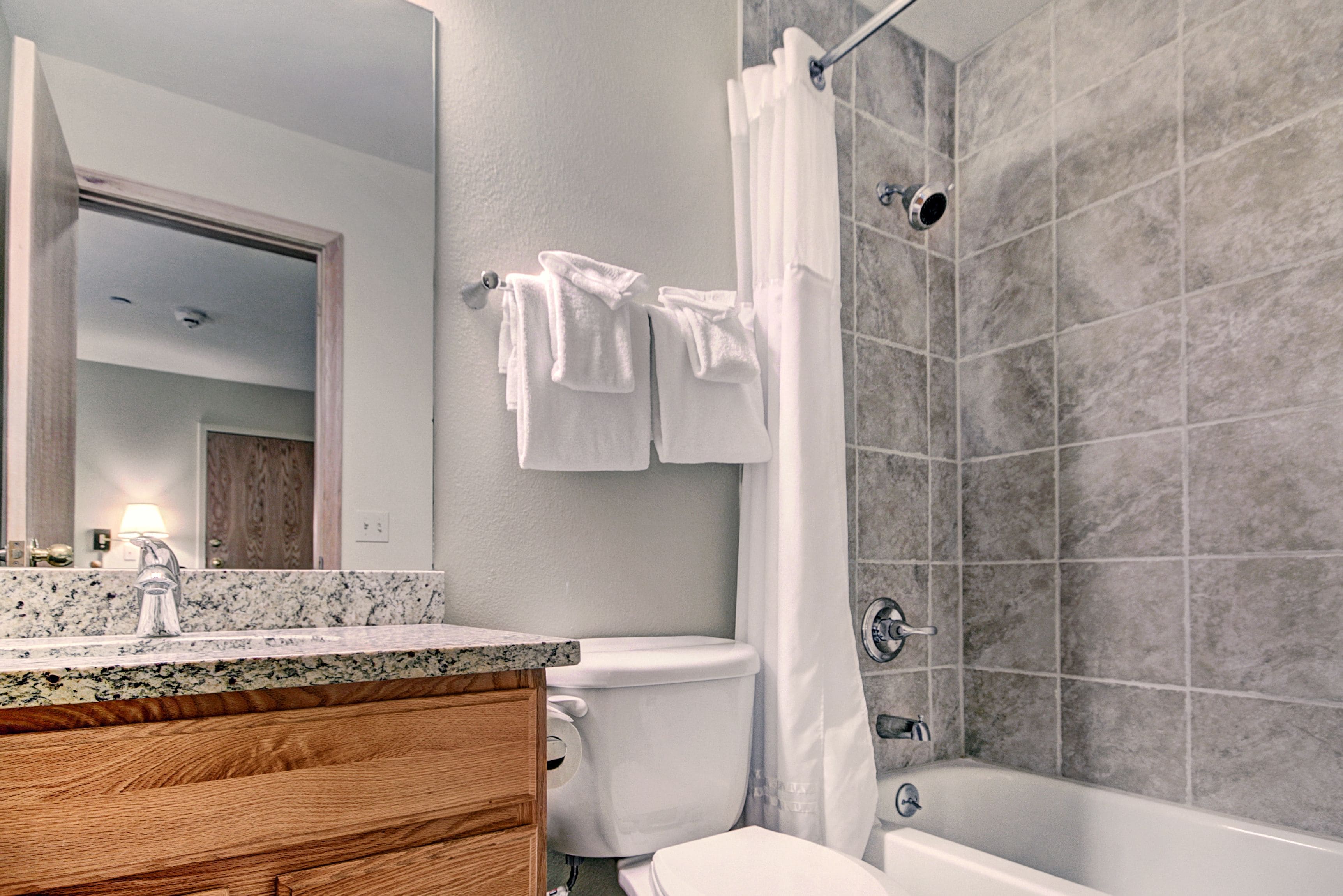 Un baño moderno con encimera de granito, tocador de madera, accesorios blancos y ducha con azulejos grises.