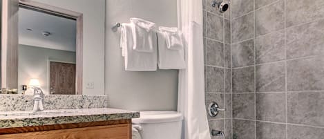 Un baño moderno con encimera de granito, tocador de madera, accesorios blancos y ducha con azulejos grises.