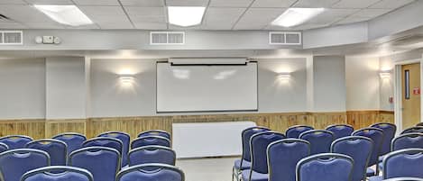 A conference room with rows of blue chairs, a white projection screen, and built-in lighting.