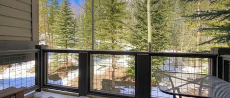 Una terraza de madera con una mesa y bancos de metal, con vista a un paisaje nevado y un bosque denso.