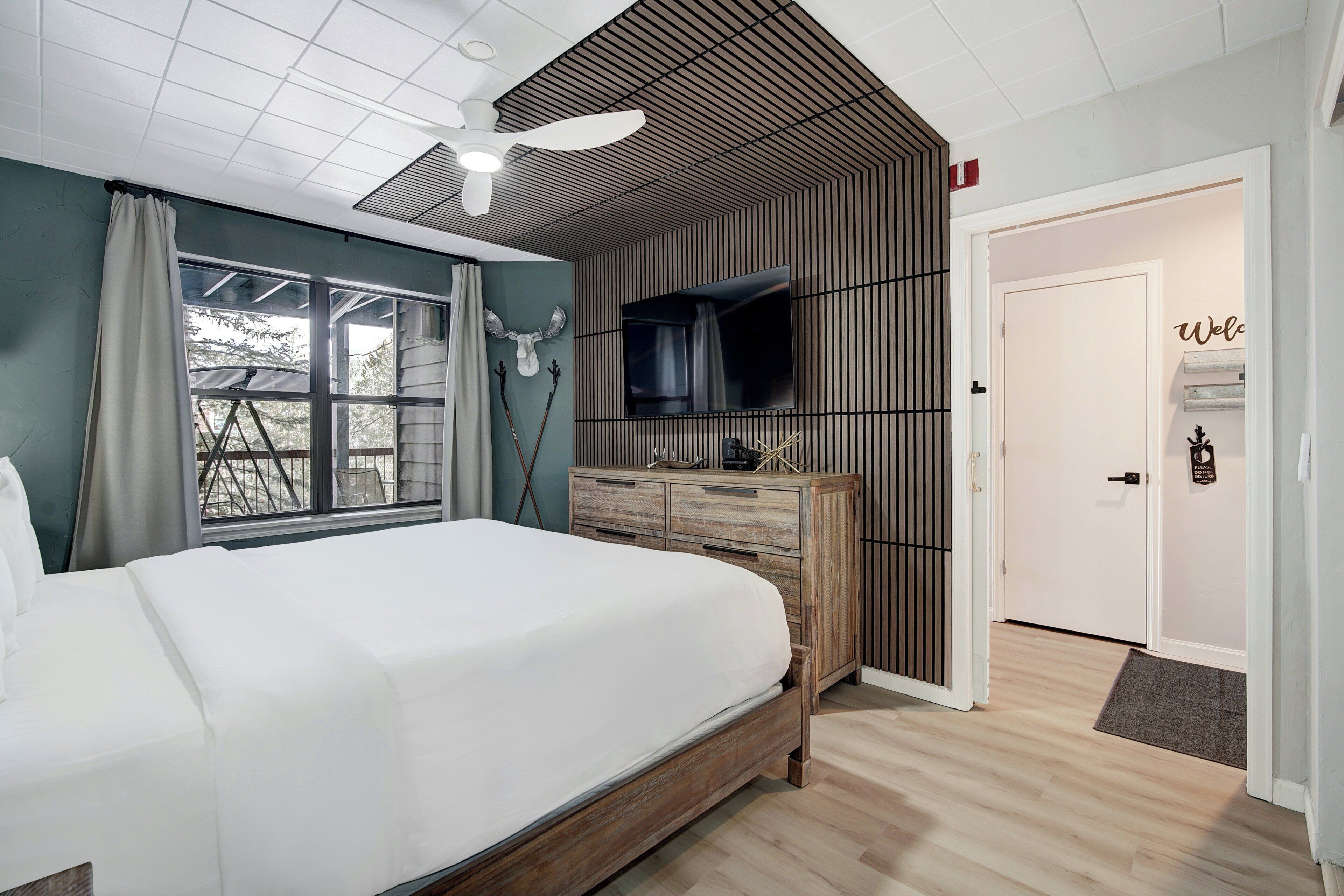A bedroom with a large bed, a TV mounted on the wall, a wooden dresser, and a ceiling fan.