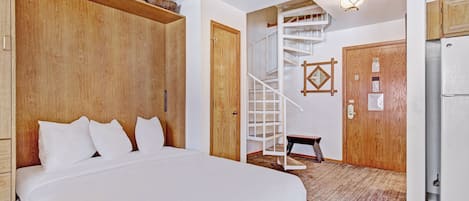 A bedroom with a large bed, wooden wardrobe, a staircase, and a refrigerator.