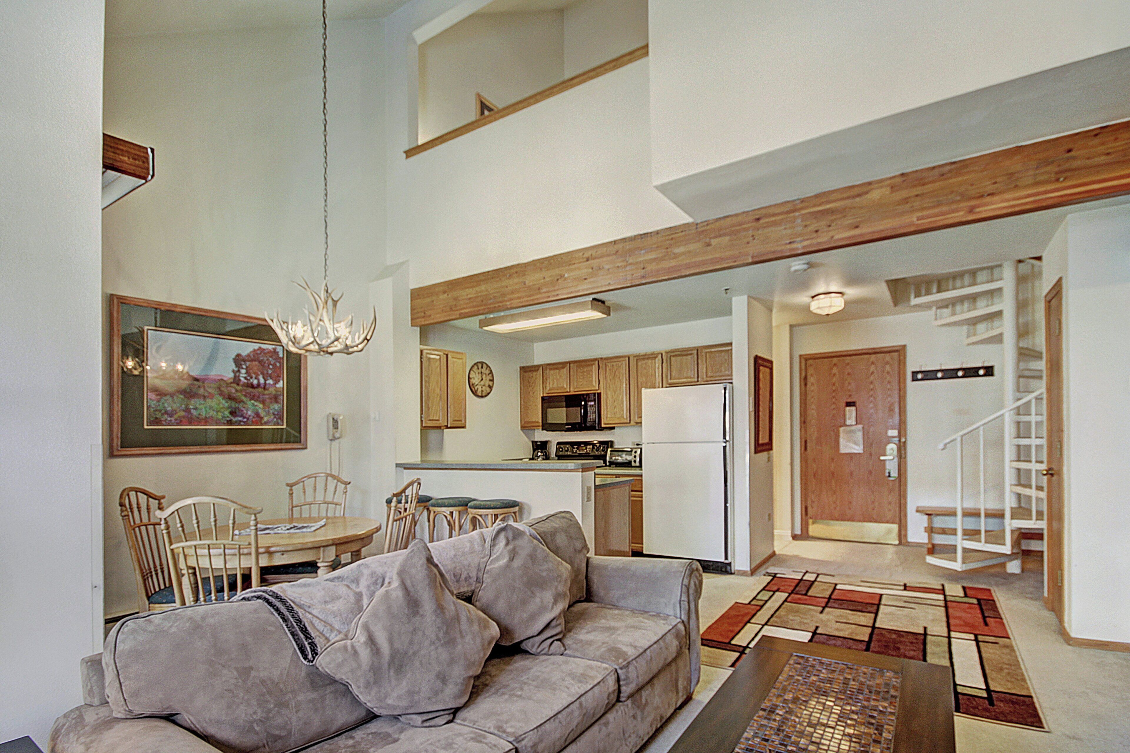 A spacious living area with a sofa, dining table, and chairs. A kitchen with wooden cabinets and appliances is visible in the background.